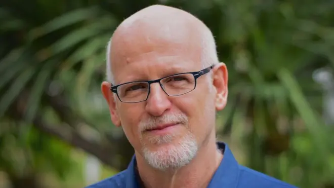 Daniel Becker é um homem de pele clara, careca, com cavanhaque e bigode brancos. Está usando um óculos retangular de aro escuro e uma camisa azul. 