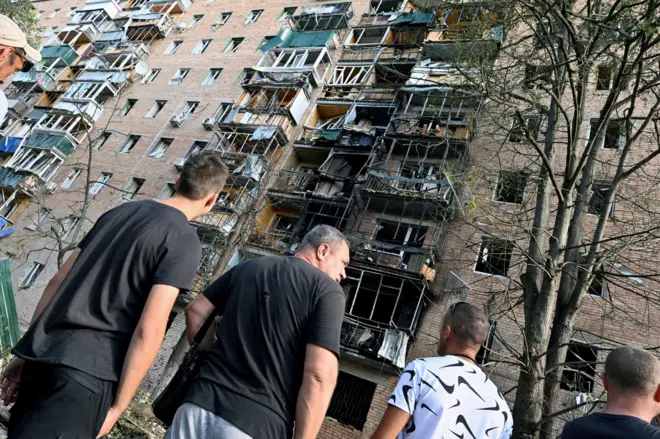 Homens parados em frente a prédio que foi atingido. Janelas quebradas e fachadas queimadas 