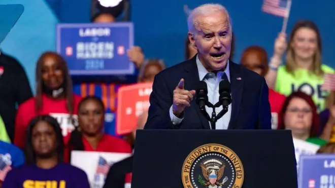 Joe Biden, um homem branco idoso de terno, discursando em frente a um grupo de jovens