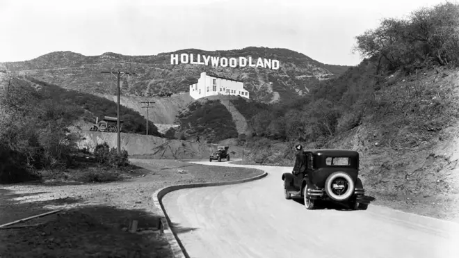 Uma placa anuncia a inauguração do conjunto habitacional Hollywoodland nas colinas de Mulholland Drive com vista para Los Angeles, Hollywood, Los Angeles, Califórnia, por volta de 1924. O edifício branco abaixo da placa é a Kanst Art Gallery, inaugurada em 1º de abril de 1924.