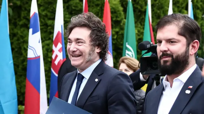 Milei e Boric caminhando e sorrindo em evento ao ar livre, onde se pode ver bandeiras de vários países ao fundo