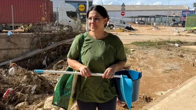 Fotografia colorida mostra mulher branca de cabelo preto carregando pá e balde em uma rua 
