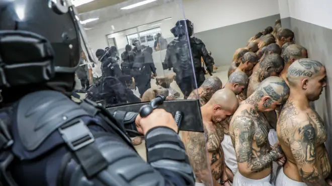 Detentos em fila, observados por policiais fortemente armados