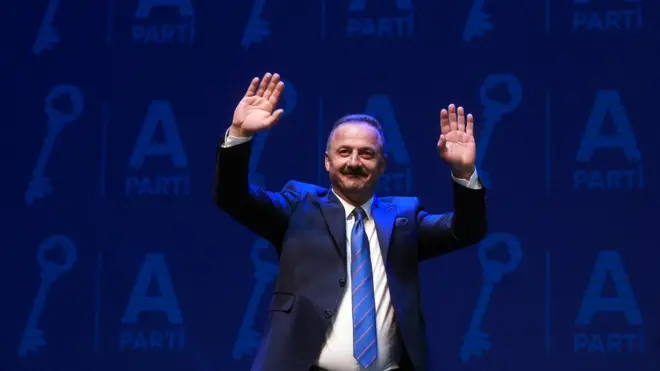 Yavuz Ağıralioğlu, partisinin logosu olan mavi bir duvarın önünde dinleyenleri selamlıyor. 