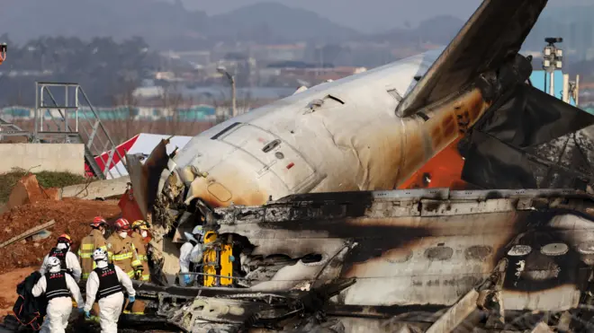 Des pompiers, certains en tenue marron, d'autres en blanc et noir, fouillent l'épave de l'avion de Jeju Air. Ils tournent le dos à la caméra. L'épave est à peine identifiable comme un avion, à part la queue.