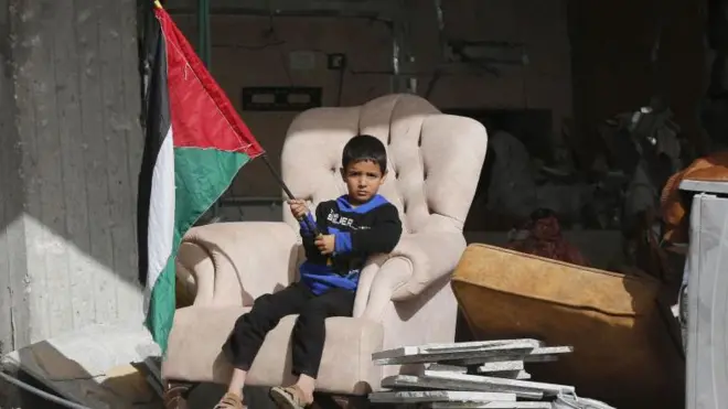 Menino segura uma bandeira palestina sentado em uma poltrona, dentro de uma casa destruída