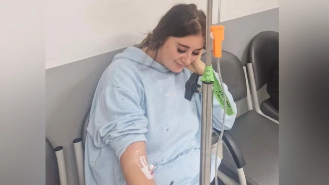 Paige Roberts sentada em uma cadeira de hospital recebendo fluidos intravenosos. Ela está vestindo uma bata de hospital azul clara e está olhando para o chão