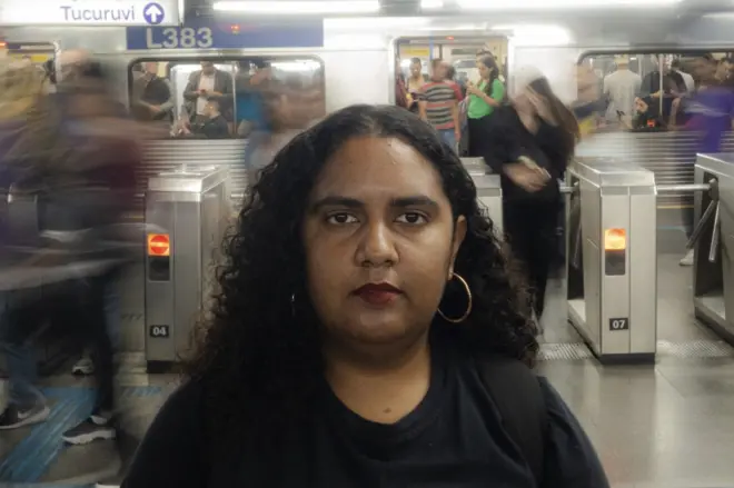 Priscila Araújo posa para foto em uma estação de metrô, onde há um vagão com pessoas ao fundo. Ela usa uma camiseta preta onde está escrito VAT - Vida Além do Trabalho