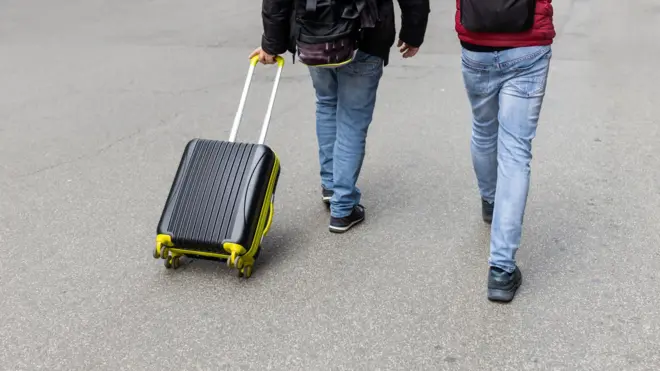 Duas pessoas caminham, de costas. Uma delas carrega uma mala de rodinhas preta