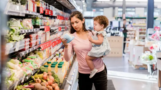 Uma mãe segurando a filha e olhando os preços dos produtos em um supermercado