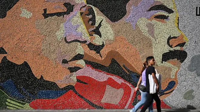 Una pareja camina ante un mural con los rostros de Hugo Chávez y Nicolás Maduro.