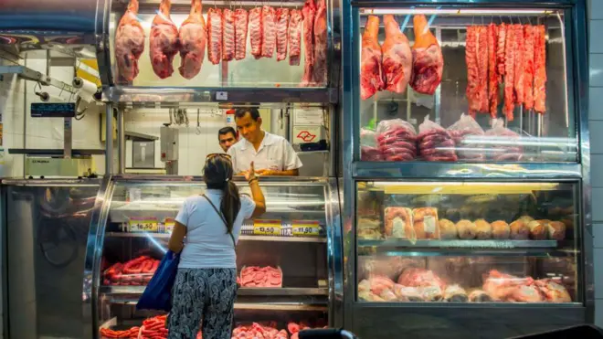 Foto mostra um homem atendendo uma mulher em um açougue. 