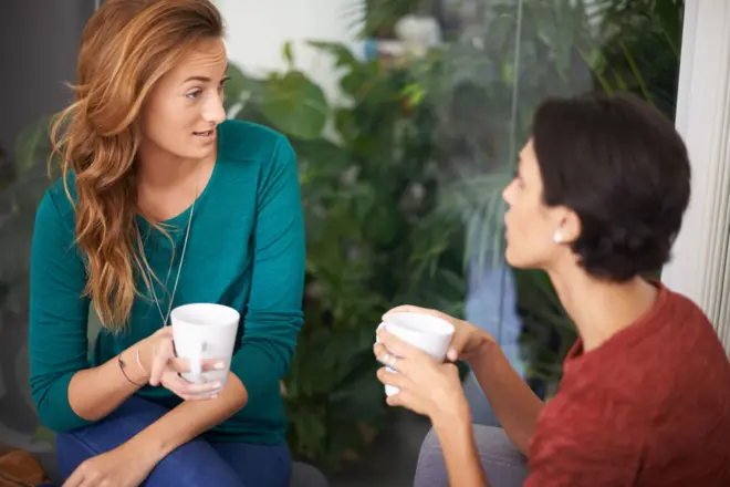 Duas mulheres brancas conversam segurando canecas nas mãos, uma delas faz cara de incredulidade