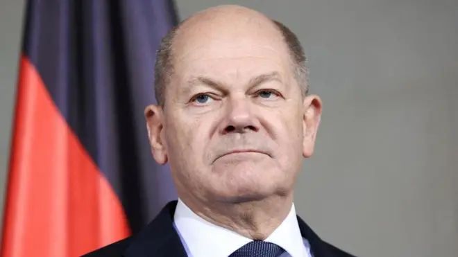 Foto do chanceler alemão Olaf Scholz, inexpressivo em frente a uma bandeira vermelha e preta