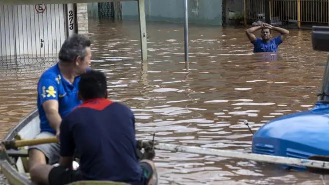 Dois homens em um barco observam um homem caminhando por uma área alagada de Porto Alegre, 5 de maio de 2024