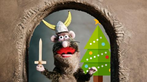 A rat wears a hat and holds a sword. A Christmas tree is in the background. Rattus Rattus from Horrible Histories.