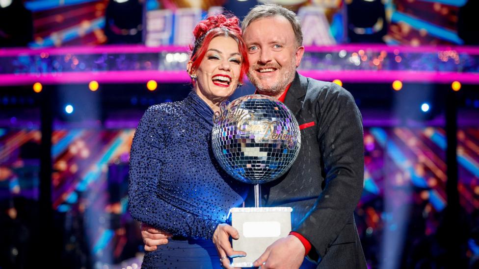 Dianne Buswell and Chris McCausland smiling and lifting the Strictly Come Dancing Glitterball trophy.