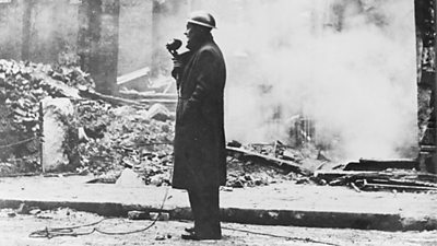 A reporter in a tin hat holding a microphone stands amid smoking rubble.