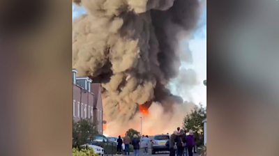 Huge fire with row of buildings in front. People stand and look towards fire.