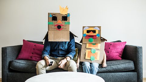 A dad and their child, with cardboard box robot masks on, sitting on a sofa