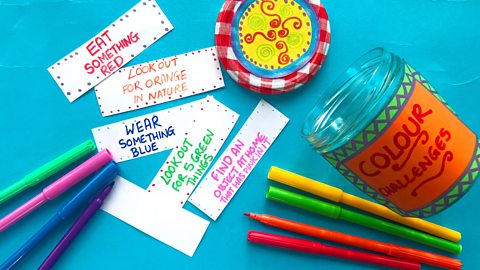 A colourful jar, a selection of colourful pens and some notes written on thin strips of paper with colour themed challenges on