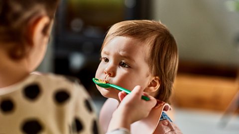 A woman spoon feeds her baby or toddler rice using a green spoon.