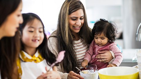 A family work together in the kitchen. In the foreground a mum and daughter look at some icing on a spoon. In the background a mum and toddler have a go at whisking a bowl.