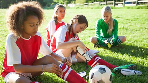 Teenage girls look disappointed after football match, adjust shin pads and reflect on game.