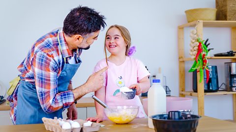 A father and his daughter are spending quality time together in the kitchen, baking a delicious treat. The father, wearing a patterned apron, is helping his young daughter, who has Down syndrome, as they mix ingredients in a bowl.