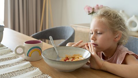 Reluctant girl sitting in front of plate with pasta in the living room and refusing to eat it.