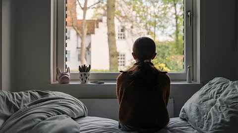 A young girl looks out a window solemnly 