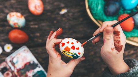 Image of someone painting an egg for Easter. There is a basket of eggs, some on the table and their hands have paint on them.