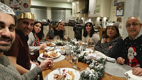 Family are gathered around a festive table wearing party crowns, eating festive food.