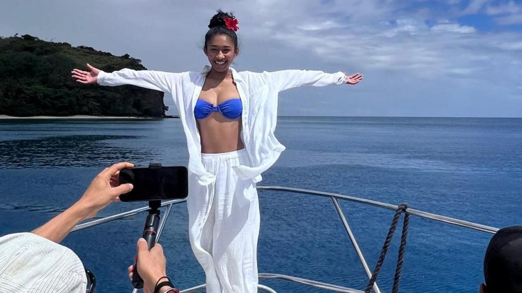 A woman wearing a white shirt and bottoms and blue bikini top poses on a boat in the sea, in front of a man picturing a picture with his phone.