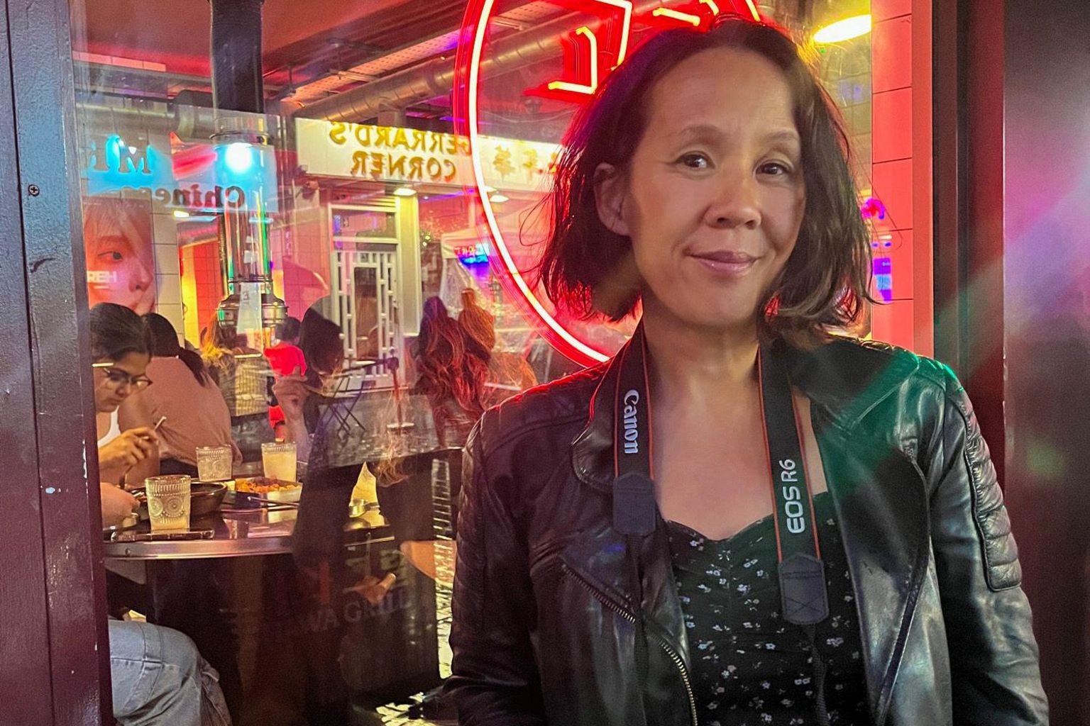 Harriet Armstrong wearing a leather jacket with a camera around her neck outside the window of a Chinese restaurant