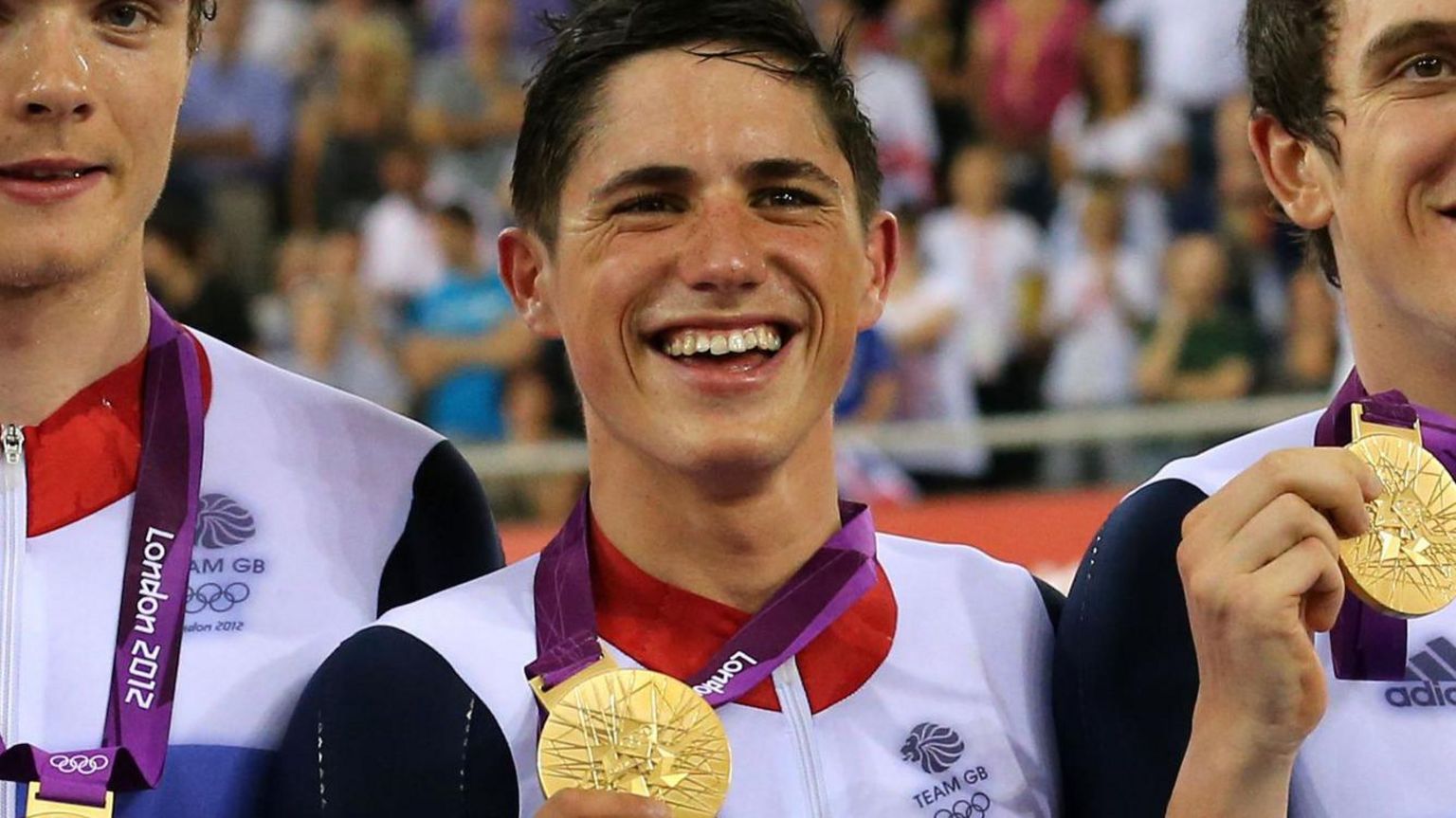 A smiling Peter Kennaugh, with short brown hair and wearing a red, white and blue top with the words 'Team GB' on it, holds his gold medal up, as he stands alongside similarly dressed Steven Burke and Geraint Thomas