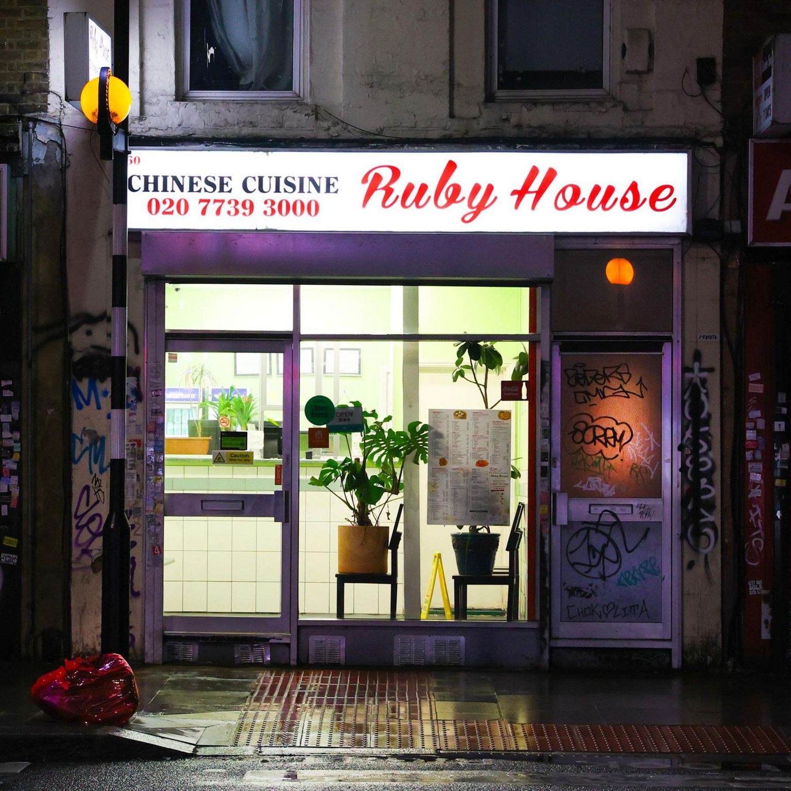 Ruby House Chhinese takeaway with plants on chairs in the window beside a door covered in graffiti