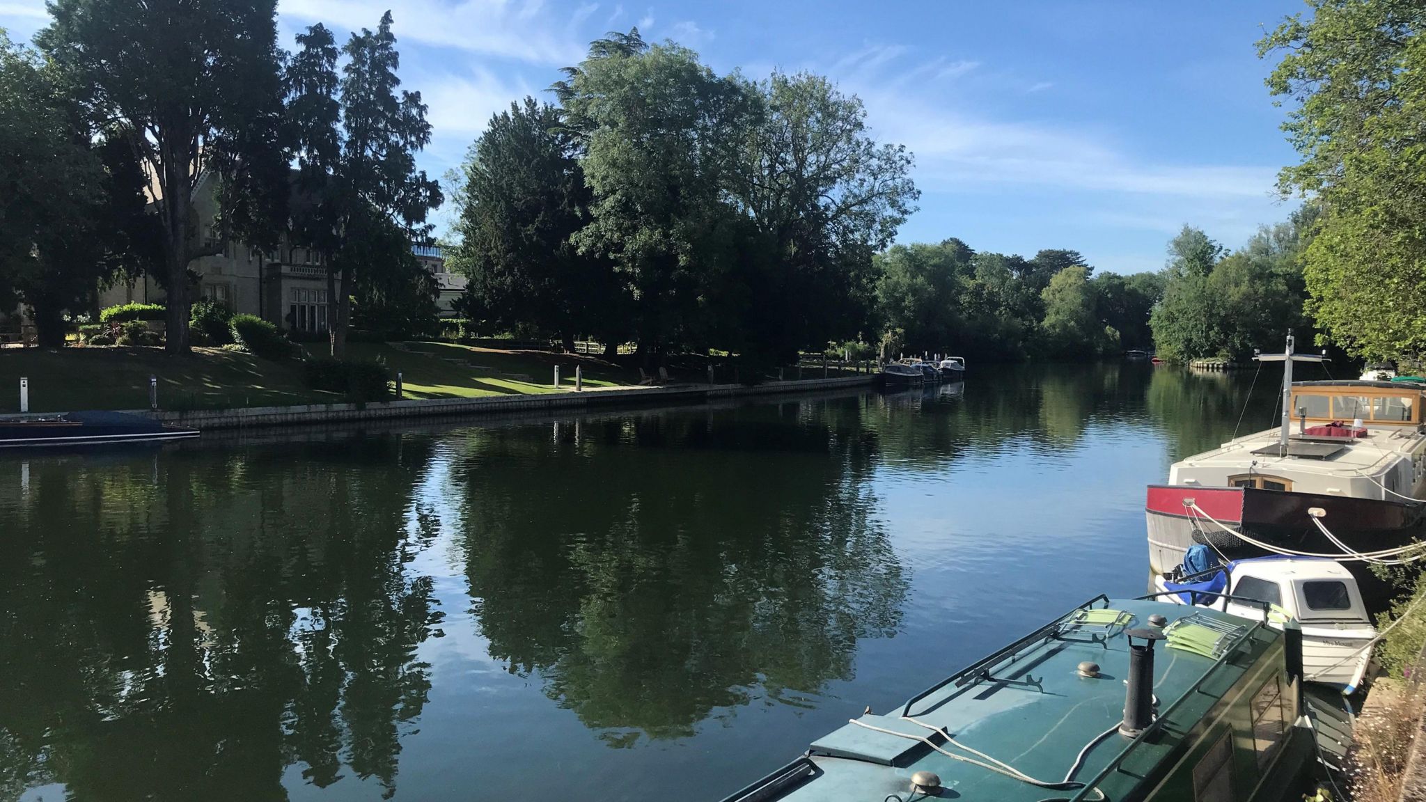 TUESADY - Boulter’s Lock at Maidenhead looking gorgeous in the sun. There are two boats moored on the right bank, a narrowboat with a green roof and a larger white narrow boat. There are trees overhanging the water and there are reflections of them in the water. On the opposite bank stands a two-storey house surrounded by trees and green grass.