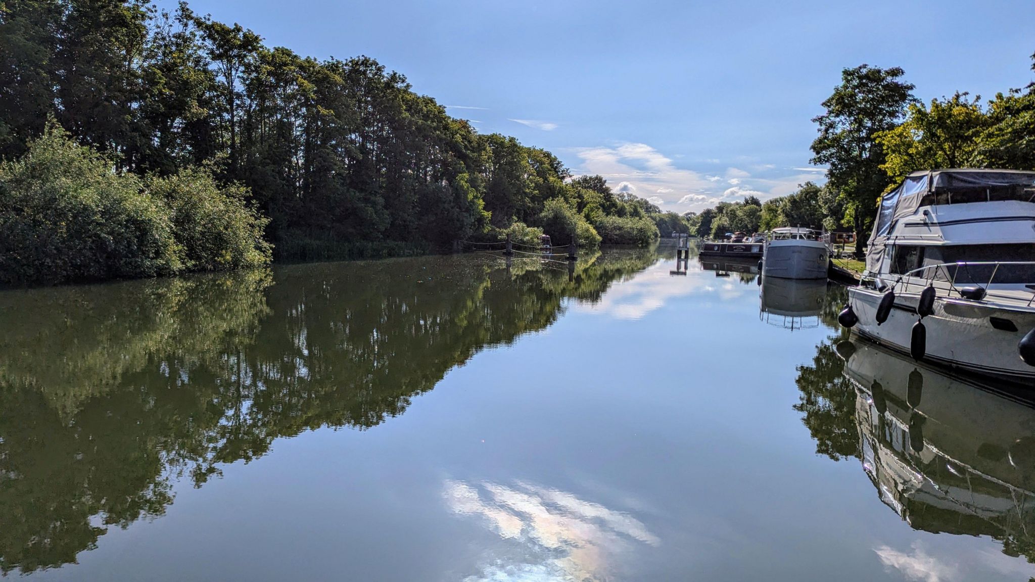 TUESDAY - A beautiful sunny day at Abingdon Lock with the sun shining on the waterway. There are green trees overhanging the left bank and a white yacht moored on the right bank. The sun and small white clouds are reflected in the still water