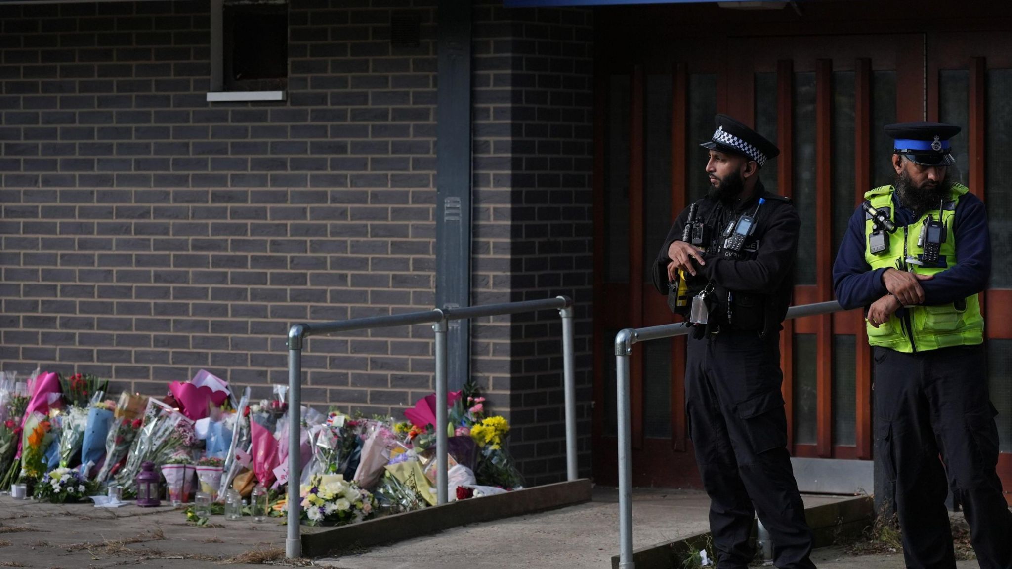 Two male police officers stand outside the Leabank flats entrance, near to the floral tributes