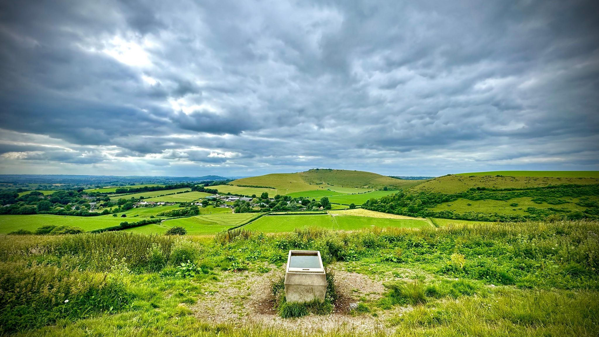 THURSDAY - The rolling green Dorset countryside is photographed from Compton Abbas. A metal water trough is in the centre of the frame, behind the countryside is dotted with trees and hedges with two hills. The sky is covered in grey skies