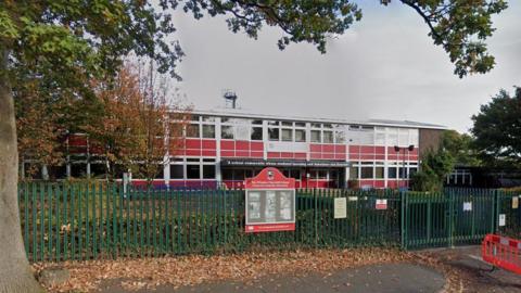 The outside of Winston Churchill School in Woking, it is surrounded by a green fence and there is red panelling on the outer walls