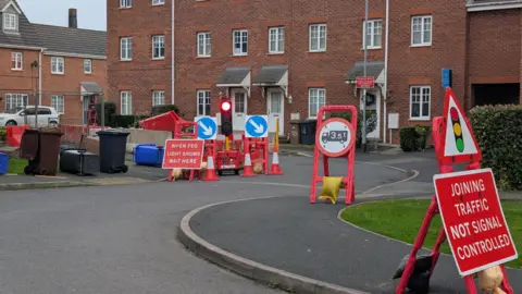 A large brick building behind a winding road with several red traffic signs, traffic cones and temporary traffic lights on it