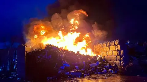 Dorchester Fire Station A large fire on a pile of metal that is contained in a constructed wall. The scene is dark.