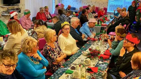 Margaret Peacock A previous Christmas Day dinner organised by Margaret Peacock in Coleraine.  Dinner guests from a mix of ages are sitting and chatting around long tables, some are wearing paper party hats, tinsel or Santa hats. 