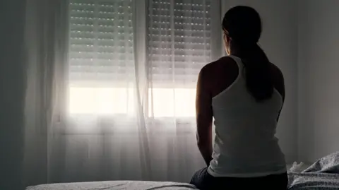 A woman with a long dark pony tail sits on a bed facing a window with the curtains and blind closed. She is wearing a white vest top. There is daylight coming through the lower part of the window and the walls are white.