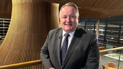 Darren Millar stood in a suit on a blacony in front of the Senedd's wooden funnel, which protrudes from the centre of the building.