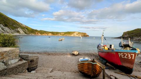 SATURDAY - Three fishing boats on the beach at Lulworth Cove. One is red and the other smaller boat is brown wood, behind is a smaller white boat that is on the shoreline. Both boats have been pulled up on the shore and behind in the cove is a pontoon and several other boats moored up. The cliffs surrounding the Cove are in the sun with green grass.