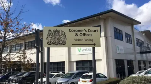 BBC A view of Suffolk Coroner's Court from the outside. It shows the entrance sign with the building sat behind it. Several cars are pictured in the car park directly outside.
