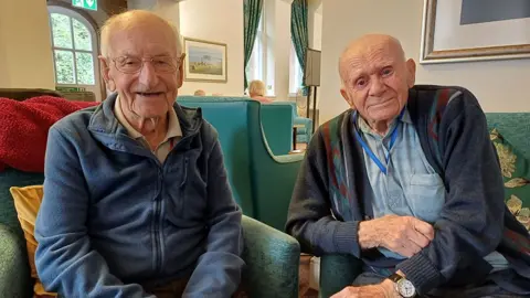 Joe Ingram and Arthur Symes looking at the camera sat in green armchairs in the Blind Veterans UK centre in Llandudno, North Wales. Joe is wearing a blue top with a zip down the middle and is balding with short white hair down the sides and wearing glasses, while Arthur has a dark blue cardigan on with a light blue shirt underneath and is bald.